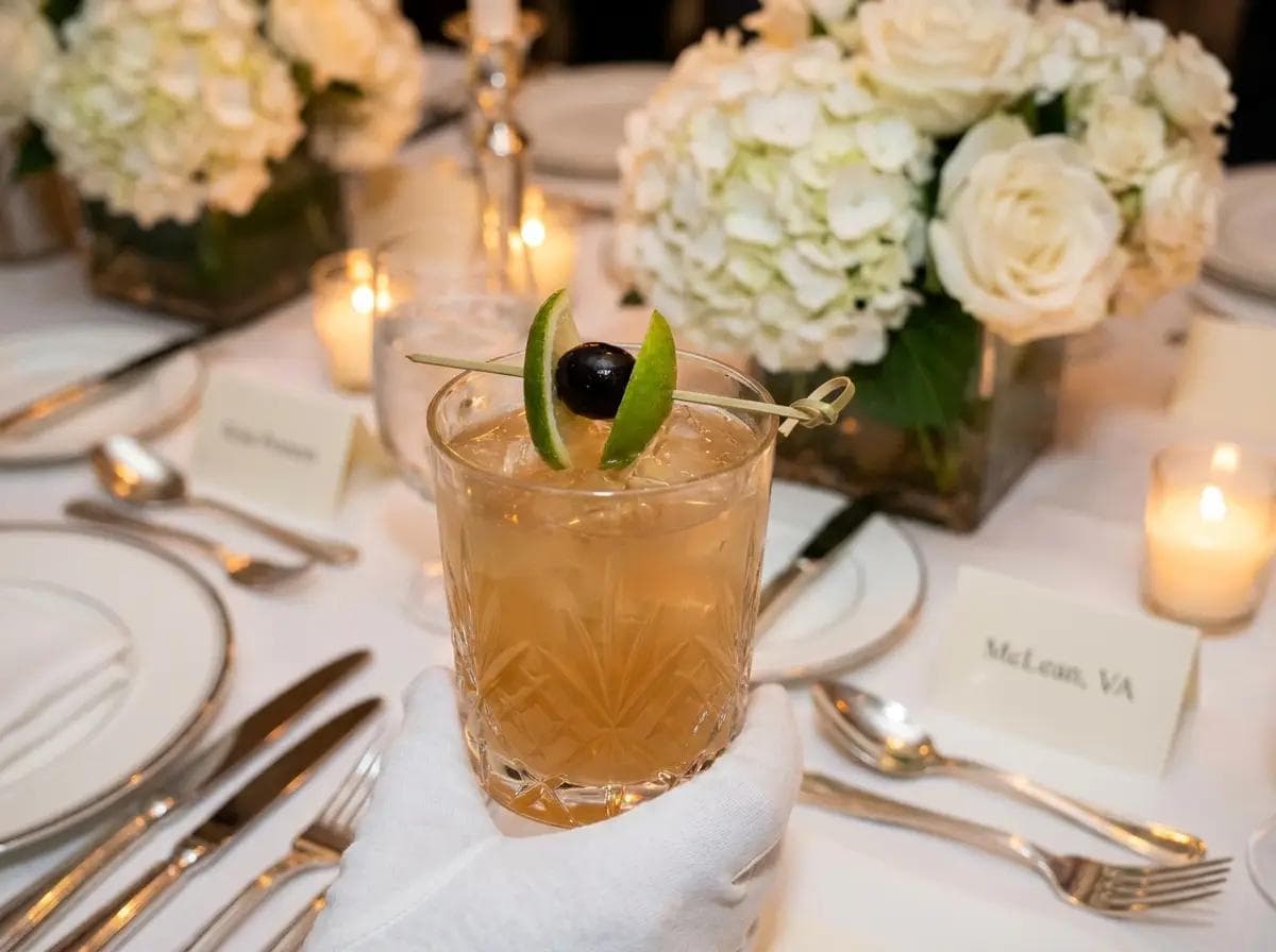 Close-up of a white-glove mocktail presentation at a McLean Virginia executive reception, crystal glassware and luxury table settings blurred behind